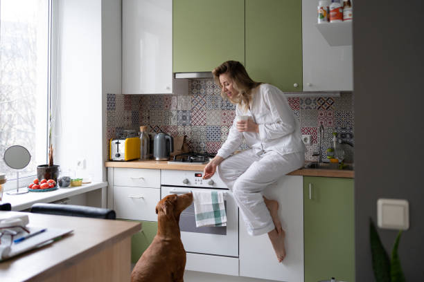 Woman in pajamas holding white mug, drinking coffee or tea in the winter morning at home, treats her beloved vizsla dog with goodies. Slow living.